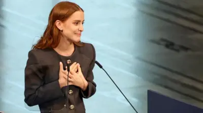 金色のボタンがついた黒い服を着たアナ・コリナ・ソサ氏が、両手を胸に当て、前方を見ている