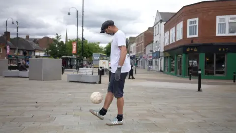 Jesse is on an almost deserted shopping street. He is wearing a white shirt and a cap and is kicking a football above his foot