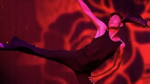 Finn on stage in a leotard and with his arms outstretched. His right leg is outstretched and he is looking off to the side against  a red swirling background.