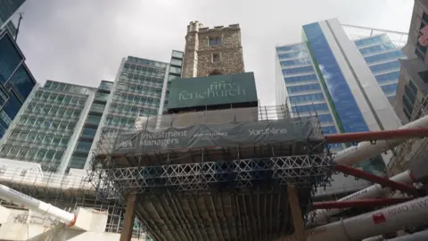 The remains of a brown bricked medieval tower is suspended on wooden slits whilst it surrounds modern skyscrapers. There are white construction pipes below the tower.