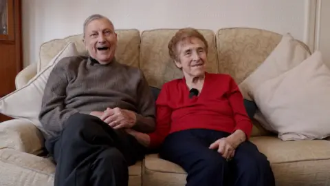 Maurice, wearing a brown jumper, and Nancy, wearing a red top, are sitting on a settee in their living room. Both are laughing.