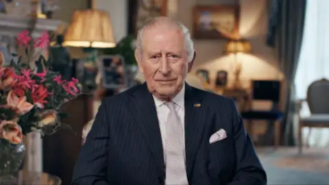 King Charles III in The Morning Room at Clarence House, London, recording a personal message. He is wearing a dark blue striped suit and pale tie, and is sitting with pink flowers on his right and there are chairs and lamps in the background. 