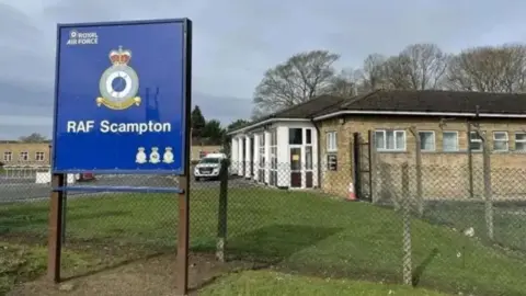 An RAF Scampton sign stands outside the main entrance to the site. There are a number of single-storey buildings in view behind a perimeter fence, along with a white vehicle.
