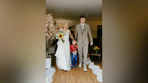 Fiona and Peter are walking together. Fiona is wearing a wedding dress and Peter is wearing a light brown suit. Their son is between them, dressed as Spiderman