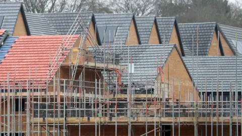 New homes under construction. The roofs of several new homes can be seen, made of red and grey slates, with scaffolding surrounding some of the homes.
