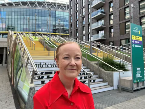A woman is smiling and looking into the camera. She has short blonde hair, and is wearing a bright red shirt, visible from the chest up. Behind her, the Spanish steps at Wembley Stadium are visible, decorated in a yellow mural, with ascending shades of yellow up the steps. The background of the steps is white, with writing at the bottom reading &#x27;And it was all yellow&#x27;. The woman is stood at the bottom left of the steps.