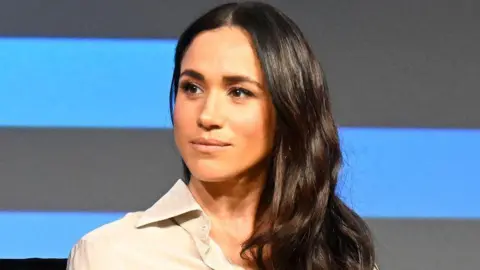 Meghan, who has long wavy brown hair, has a neutral expression as she looks on during a talk on stage