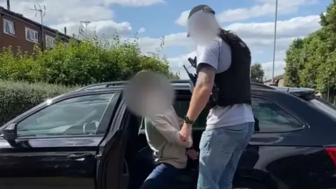 A plain-clothes police officer guides an arrested suspect into the back of a car. Both men&#x27;s faces are blurred