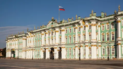 Hermitage Museum photograph showing the entire building. It is mint green and white with gold detailing. There is a pole with a Russian flag raised from the front centre of the roof