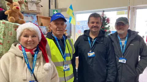 A woman wearing a white jumper and a Santa hat, stood next to three men, all of them are wearing tags which say &#x27;Hereford Help for Ukraine&#x27;. Behind them are shoeboxes wrapped in Christmas paper. 