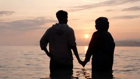 Couple stand in the sea with the sun in the background
