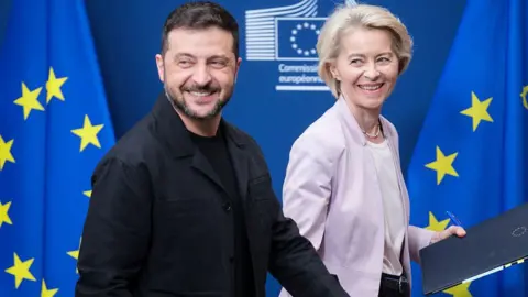 President of Ukraine Volodymyr Zelenskyy (L) and the EU Commission President Ursula von der Leyen (R) walk in front of blue and yellow-starred European Union flags