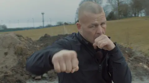 Gary Fox in a black tracksuit throwing a punch towards the screen. He is bald and is stood in a field. 
