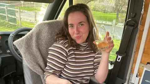 A woman with mid-length brown hair sat inside a van drinking a glass of juice, eating her breakfast 