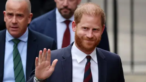 Prince Harry, wearing a dark suit and striped tie, waving to cameras
