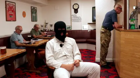 A man in a balaclava is sitting in a bar, with men drinking pints behind him.