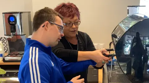 Kyle Hobbs wears a blue fleece jacket with three white stripes along the arm. He has short brown hair and wears glasses. He is being taught how to operate a coffee machine by an older woman who has short red hair and glasses. 