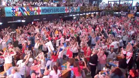  A large crowd in red and white shirts stands and cheers in celebration at Boxpark in Wembley.