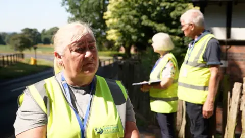 A woman with grey hair tied back in a short sleeve grey t-shirt and a hi-vis jacket with two people behind her at a roadside
