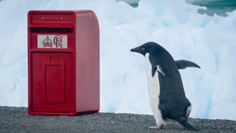 A red post box bearing the crest of King Charles III is sitting on a rocky outcrop with snow in the background. A penguin is standing beside the box.