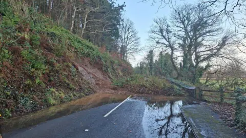 A steep bank on the left of the road, there is mud and foliage blocking the entire carriageway and there is a large puddle in the foreground.
