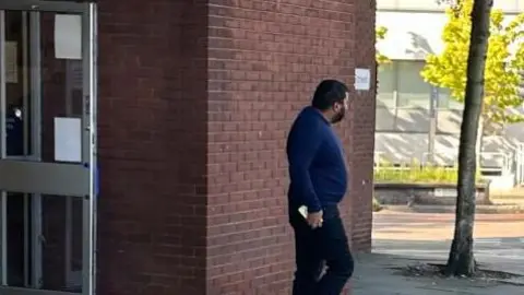 James Ayoola outside Ipswich Magistrates&#x27; Court wearing a blue jumper, dark trousers. He has short dark hair and a dark beard. He is looking away from the camera. 
