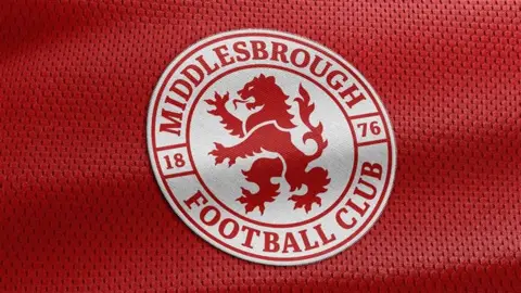 The new badge which is a white circle with a red lion in the centre of it. &#x27;Middlesbrough Football Club&#x27; is written in red letters around the circle, with the year 1876. The badge is sitting on what looks like a red sports shirt.