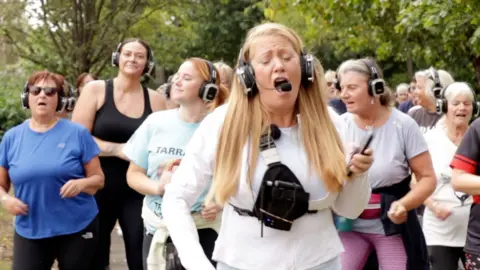 A group of women are dancing in front of trees. They are all wearing headphones.