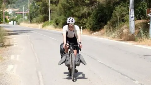 A picture of Duncan on a bike, cycling on a large path. It is a black bike, with a man wearing a white shirt and white helmet. There is grassland and poles either side of the road.